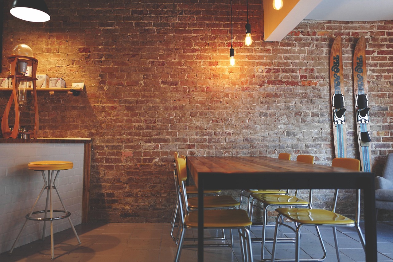 A long wooden table with yellow chairs against a brick wall under hanging bulbs